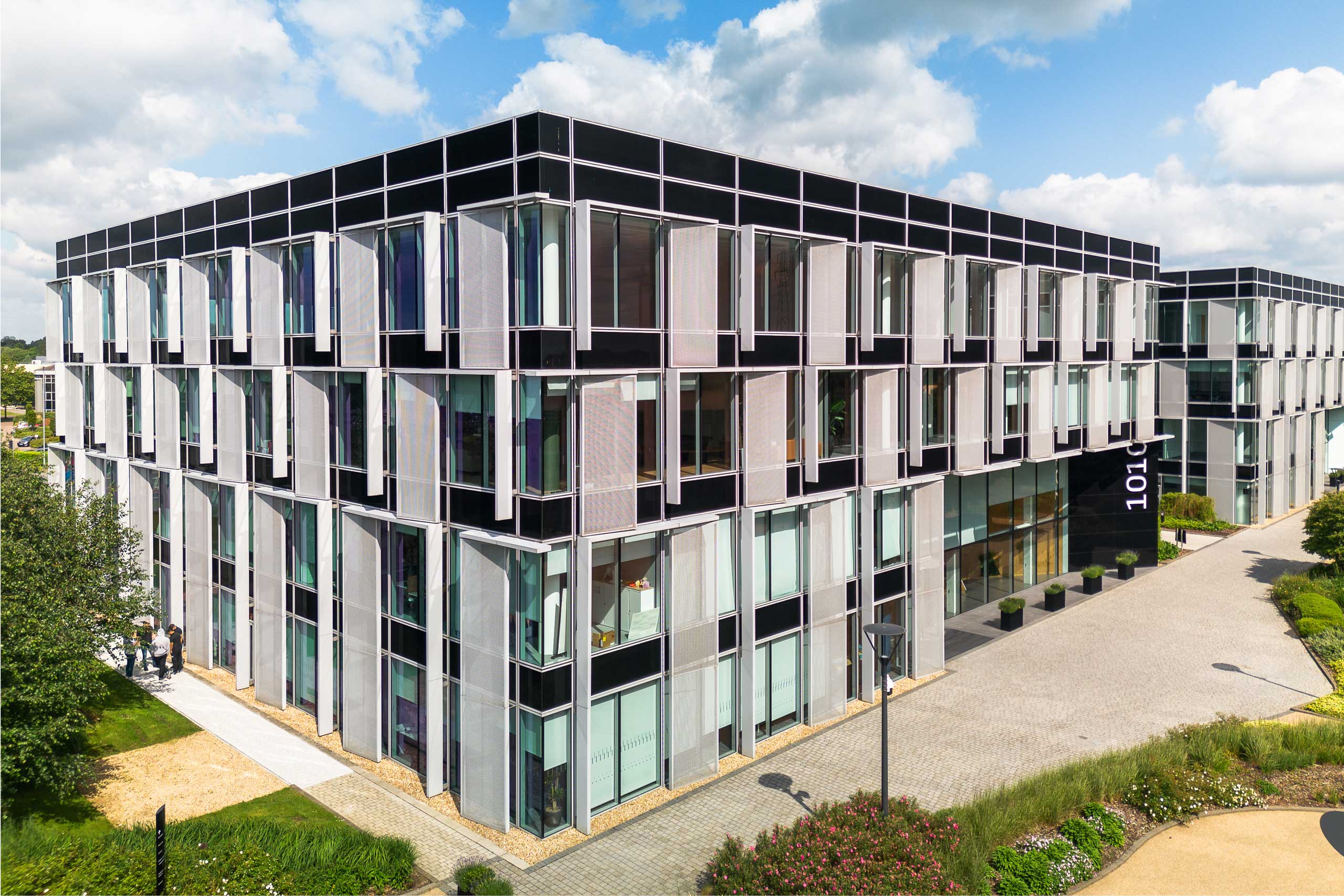 Modern office building with large glass windows, black and white panelling, and landscaped pathways under a partly cloudy sky.