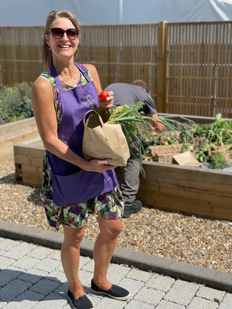A woman in a purple apron stands outdoors holding a paper bag of vegetables and a tomato. A person in the background works in a raised garden bed.