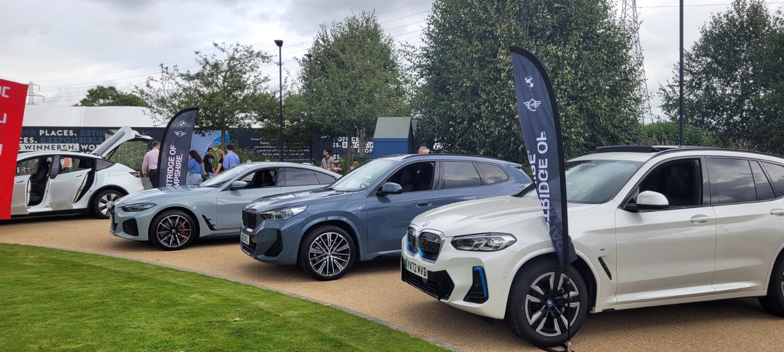 Three BMW electric vehicles are displayed outdoors on a path, with informational banners beside them and people observing in the background.