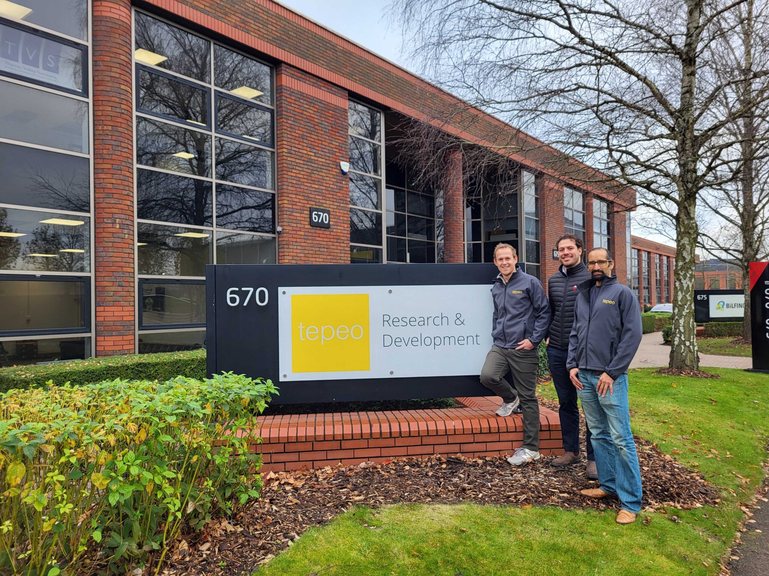 Three people stand next to a sign reading tepeo Research & Development outside a brick office building with the number 670.