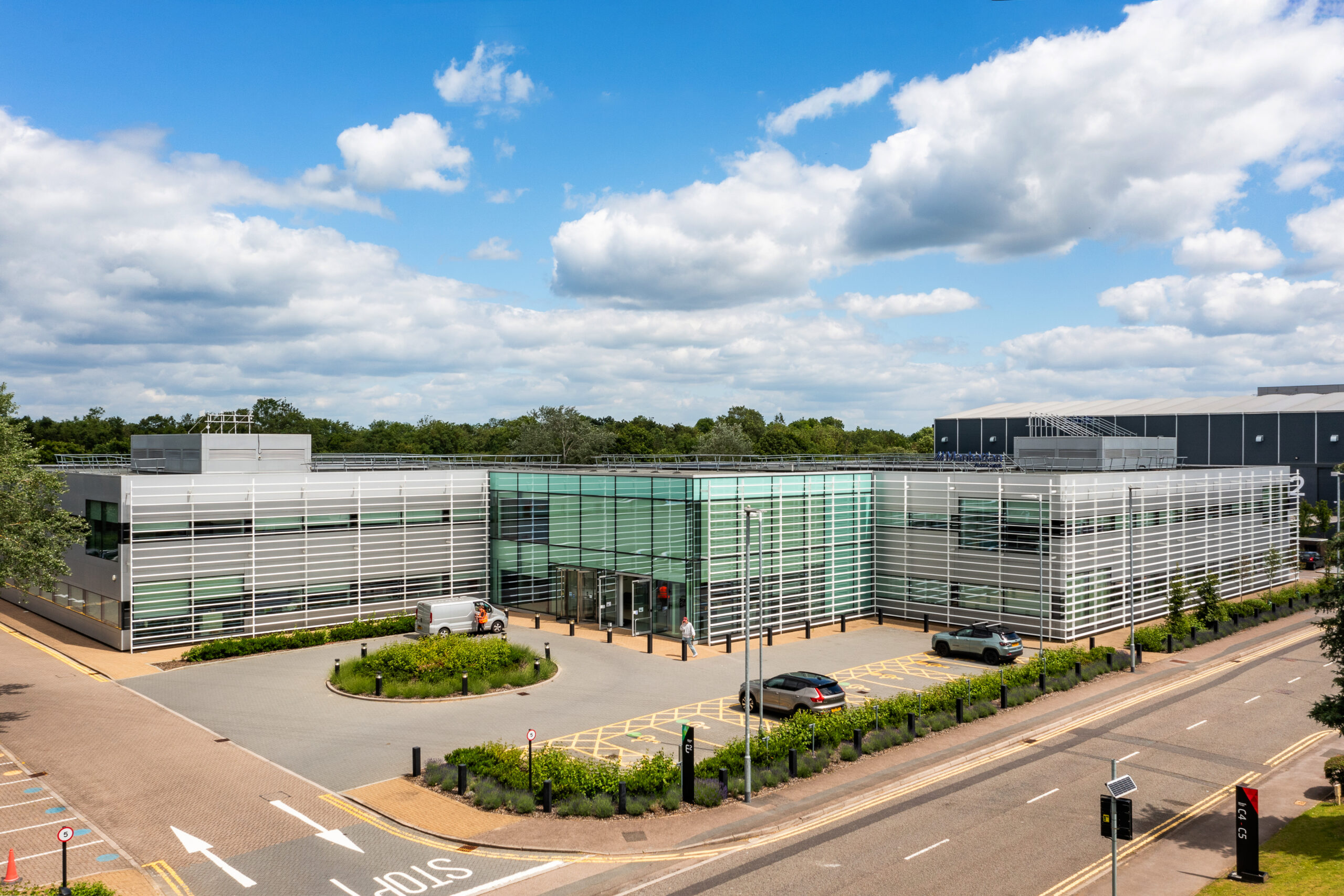 Modern office building with glass and metal exterior, surrounded by greenery and parking bays, under a partly cloudy sky.