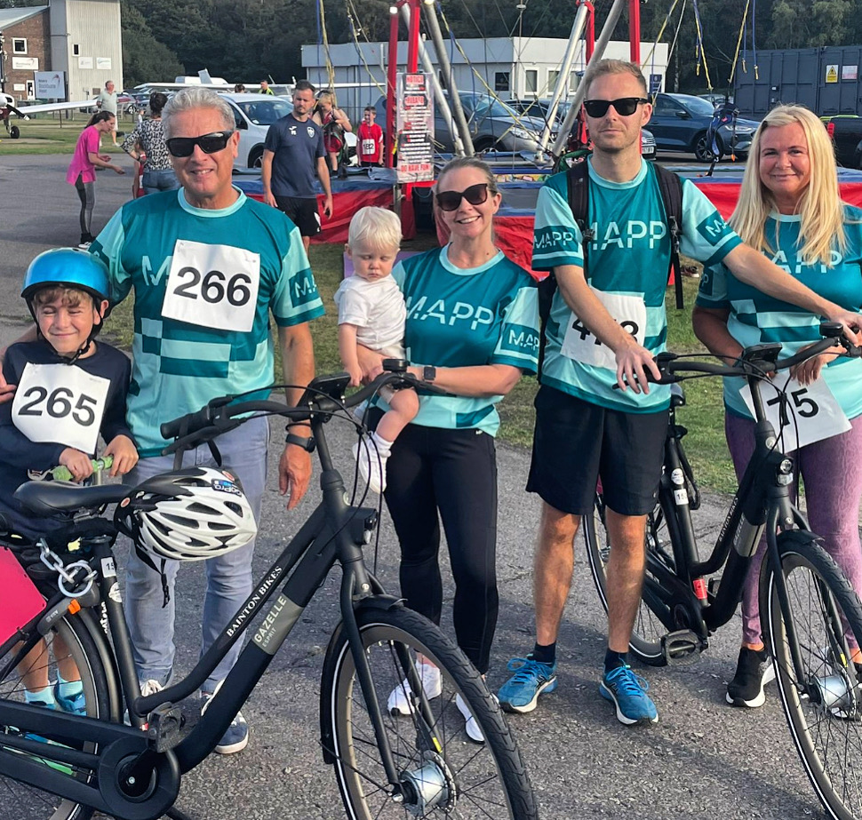 Five adults and one child stand outdoors with bicycles, wearing numbered race bibs and matching teal shirts, posing for a group photo at an event.