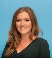 Woman with long light brown hair, wearing a dark green jumper, smiling in front of a plain blue background.