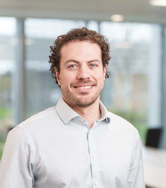 A man with short curly hair and a beard, wearing a light patterned shirt, stands indoors in an office setting with large windows in the background.
