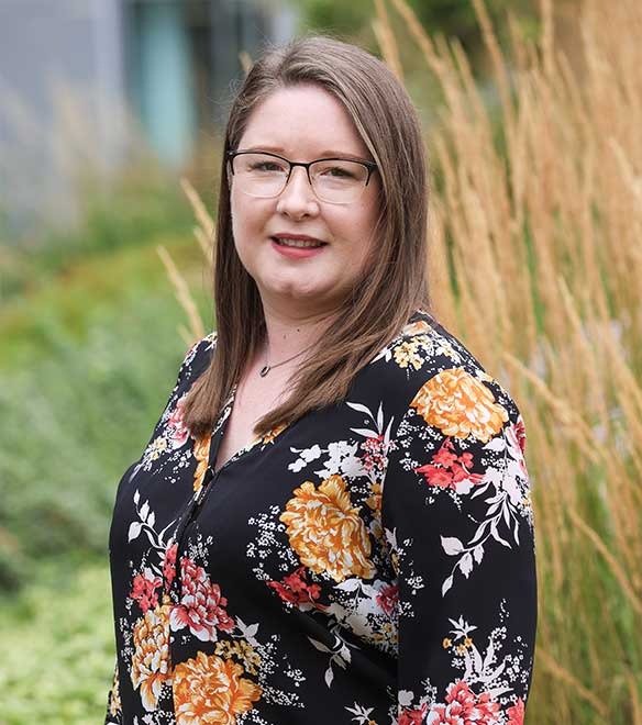 A woman with long brown hair and glasses stands outdoors in front of tall grass, wearing a black floral blouse and smiling at the camera.