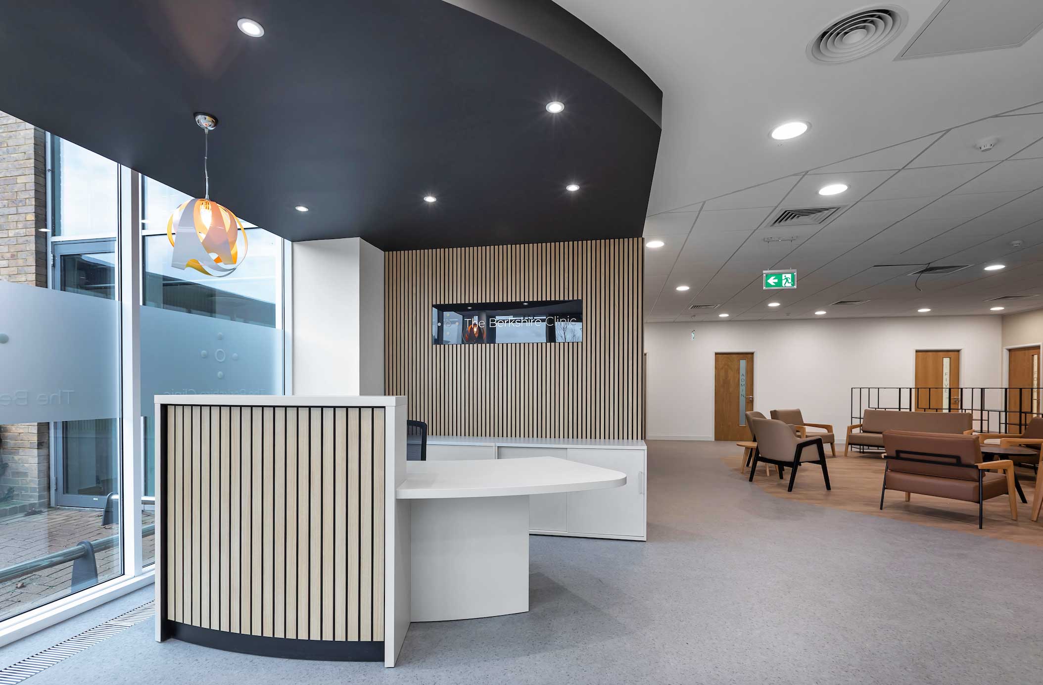 Modern office reception area with a white curved desk, wooden panelling, brown lounge chairs, and a wall-mounted TV screen. Large windows allow natural light into the space.