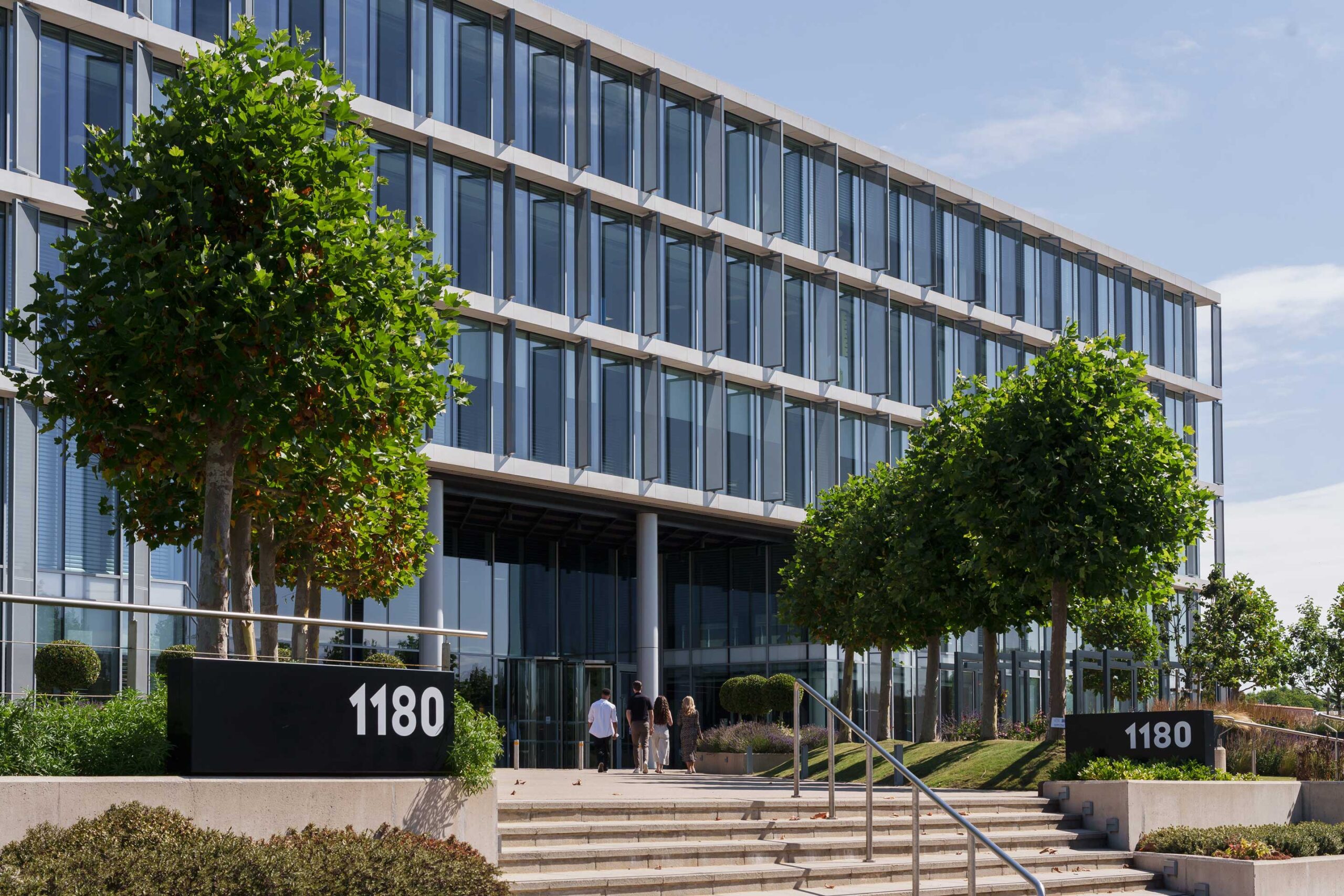 Modern office building with glass windows, two large trees by the entrance, and the number 1180 displayed on signs beside the steps.