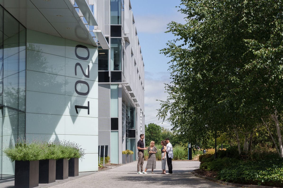 Four people stand talking outside a modern building with the number 10200 on its exterior, surrounded by greenery and trees on a sunny day.