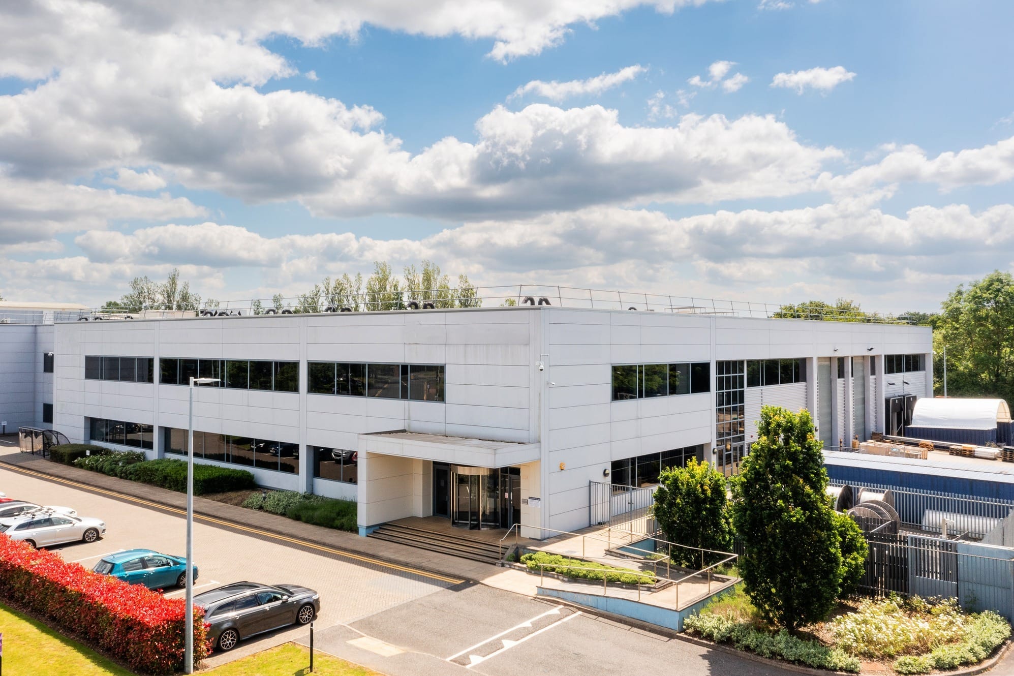 A modern, white industrial or office building with large windows, surrounded by parked cars, trees, and a clear sky with scattered clouds.