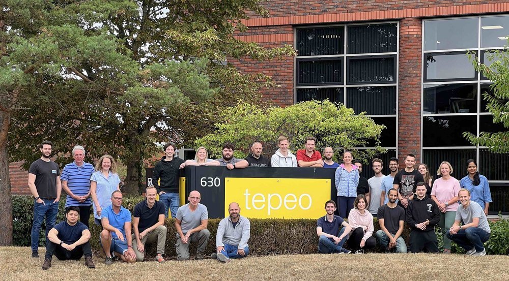 A group of around 30 people posing in front of a brick building and a large yellow sign that reads tepeo 630, with trees and bushes in the background.