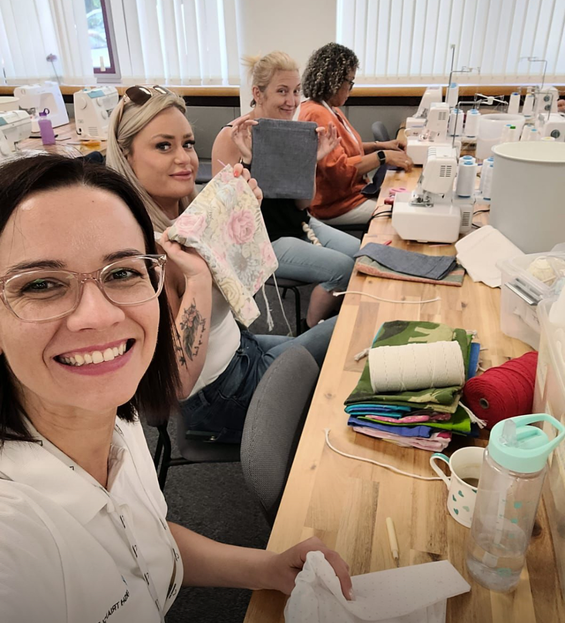 Four women sit at a table with sewing machines and fabric, smiling at the camera. One woman holds up pieces of patterned cloth. Various sewing materials are on the table.