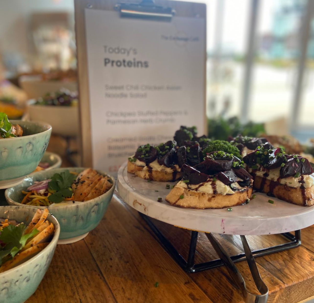 A display of salads in ceramic bowls and bruschetta topped with greens on a marble platter with a protein menu in the background.