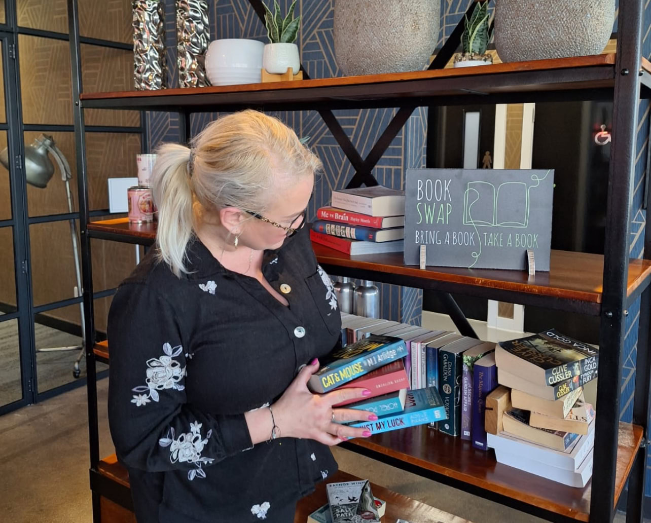 A woman stands in front of a bookshelf labelled Book Swap, holding several books and looking at the shelves filled with various titles.
