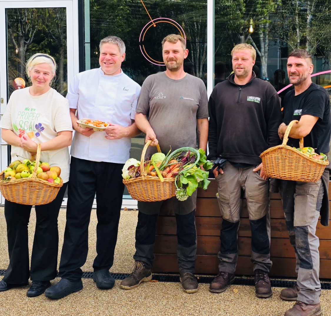 Five people stand outside a building holding baskets of assorted vegetables and fruit, with one person in a chef’s coat holding a plate of food.