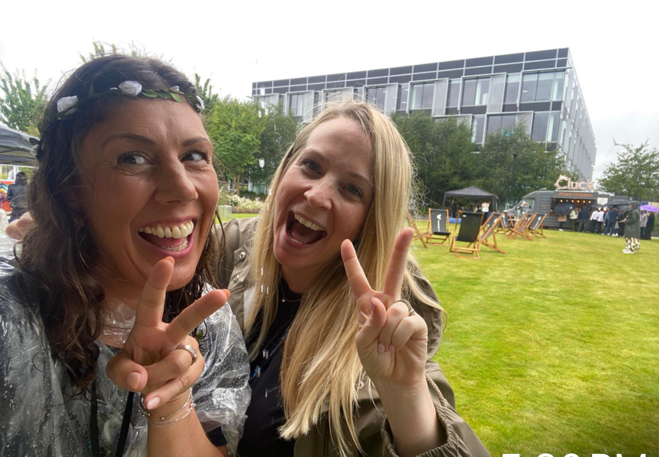 Two women smiling and making peace signs pose for a photo outdoors on a grassy lawn with deckchairs and a modern building in the background.
