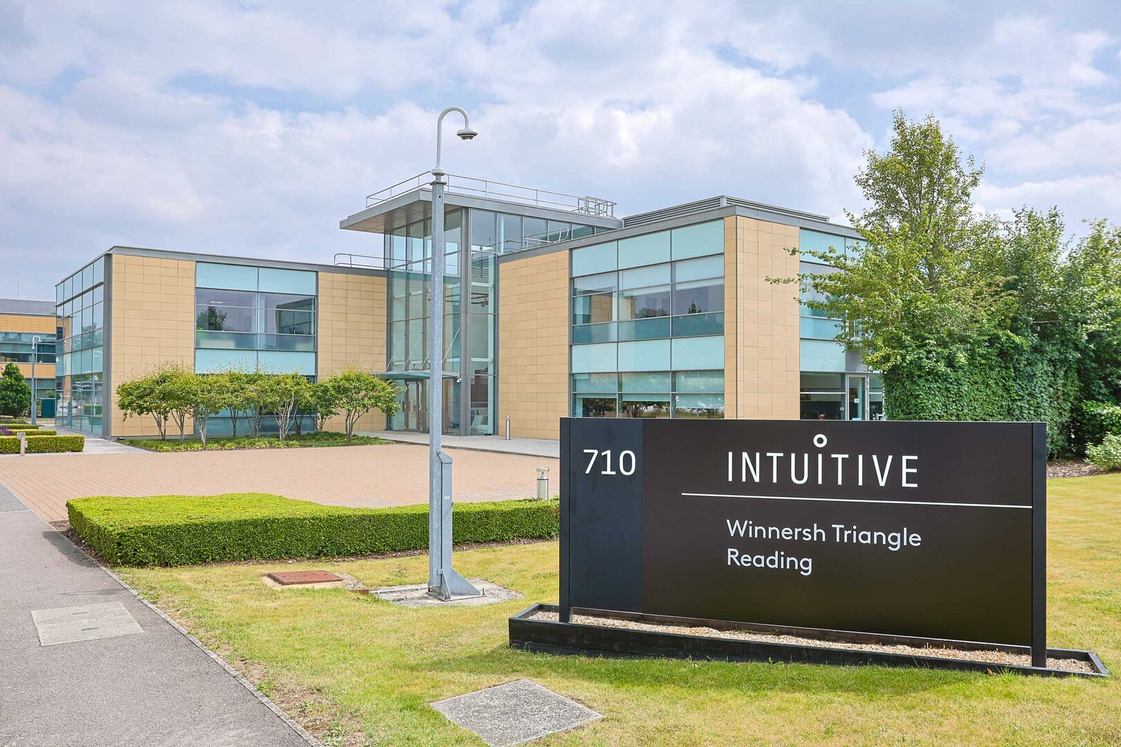 Modern office building with glass and beige walls, surrounded by landscaped greenery; a black sign in front reads 710 INTUITIVE Winnersh Triangle Reading.