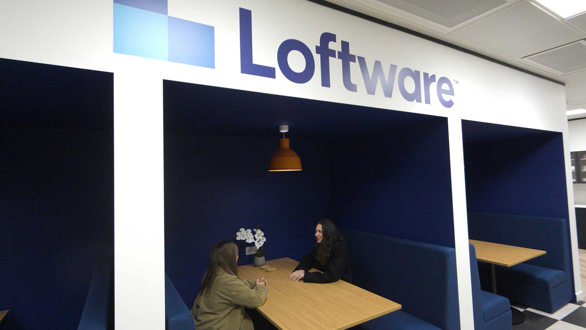 Two people sit and talk at a wooden table in a blue booth beneath a sign that reads Loftware inside a modern office space.