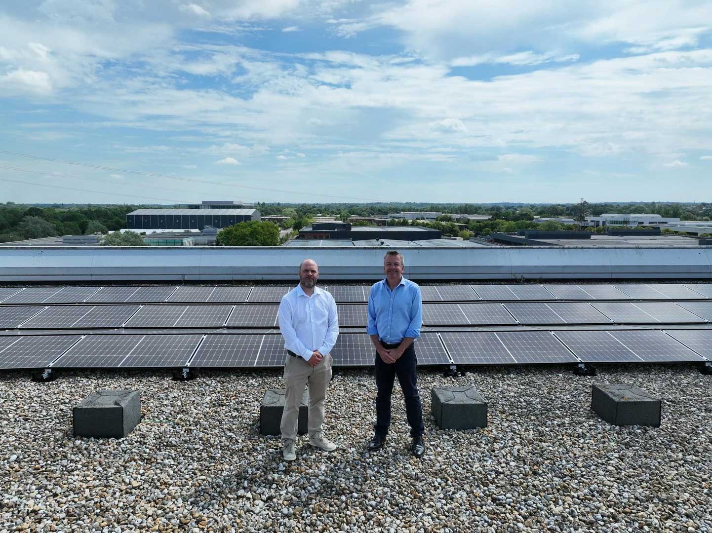 Two men stand on a gravelled rooftop in front of solar panels, with industrial buildings and trees visible in the background under a partly cloudy sky.