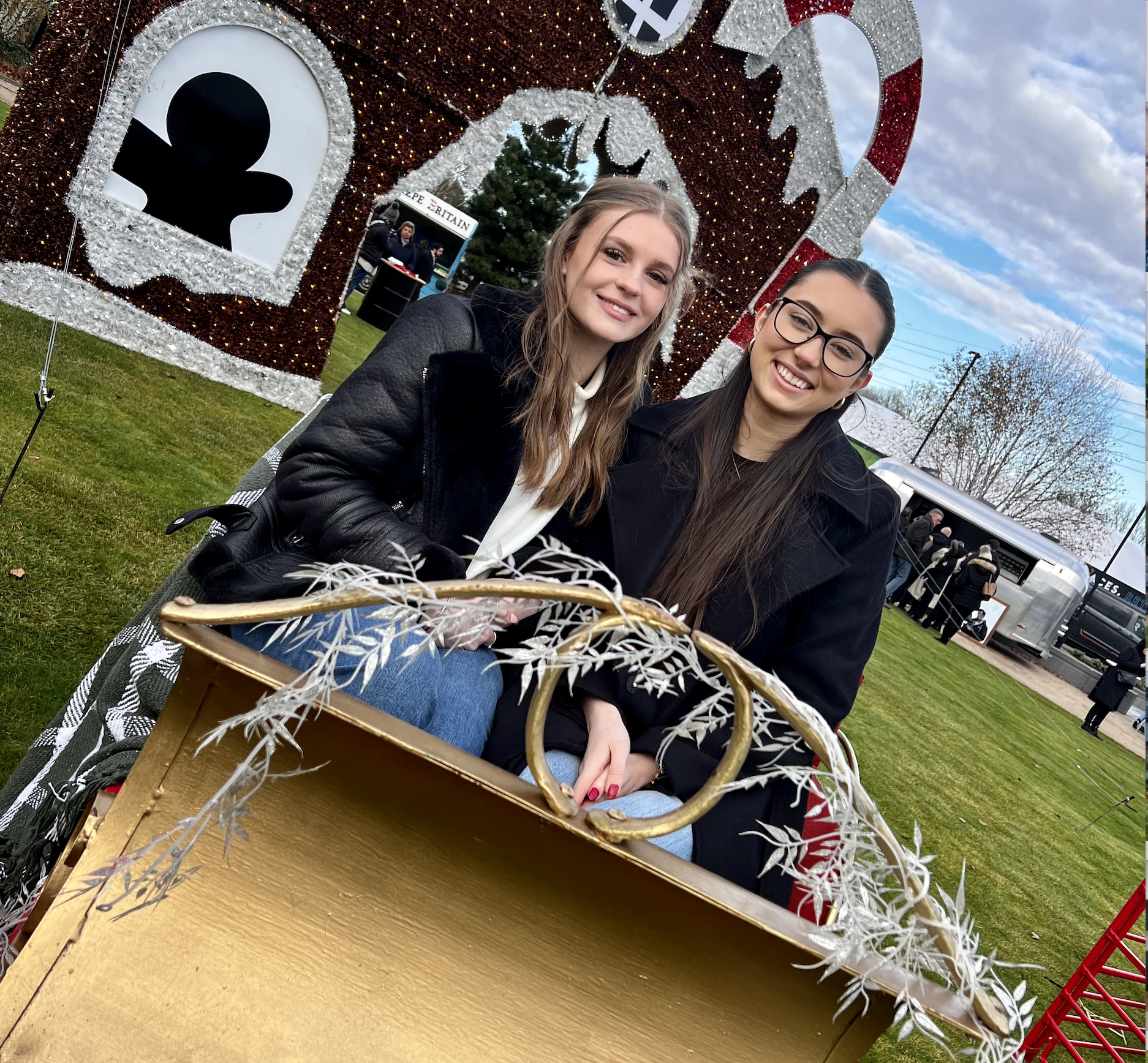 Two young women sit in a decorative gold sleigh outdoors at a holiday-themed event, with festive decorations and people in the background.