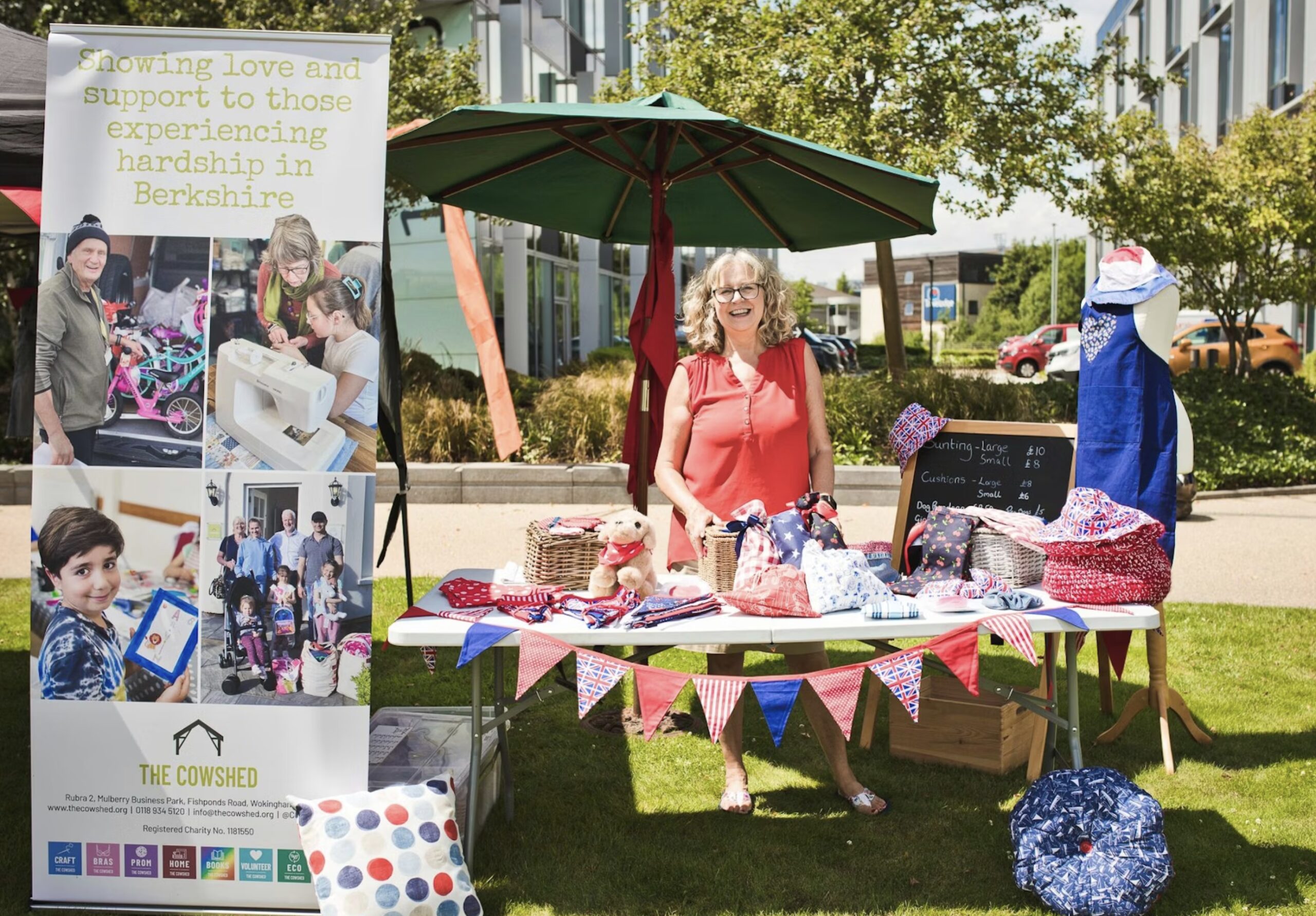 A woman stands behind a craft stall decorated with bunting at an outdoor market, with a banner for a Berkshire charity displayed on the left.