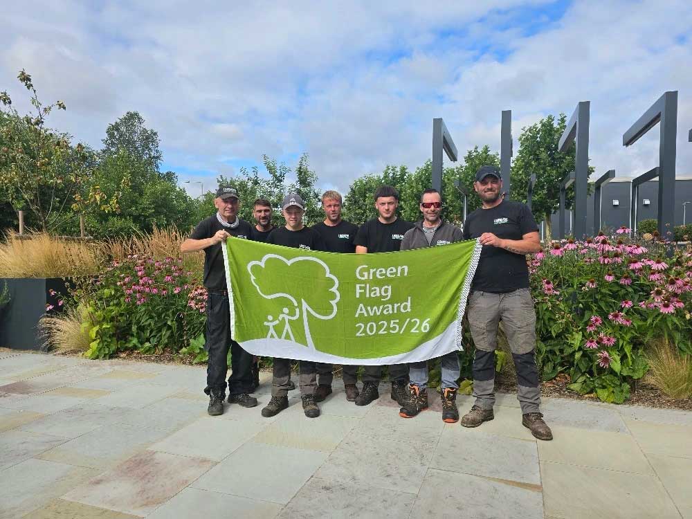 Six people stand outdoors in front of flowers and greenery, holding a green flag that reads Green Flag Award 2025/26.